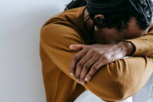 Person sitting with head down, arms folded on their knee, wearing a mustard-brown sweater (conveys fatigue or sadness).】【Close-up of someone resting their head on crossed arms on their knee, in a brown sweater.】【Individual leaning forward, head bowed onto their arms across a knee, wearing a tan knit sweater.】【A reflective moment: person with dreadlocks resting on their arms on their knee in a brown sweater.】【Person in a brown sweater sits with head tucked into their arms, suggesting tiredness or pensiveness.