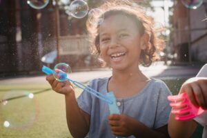 Smiling girl blowing bubbles outdoors with a blue bubble wand in hand and bubbles floating around her.