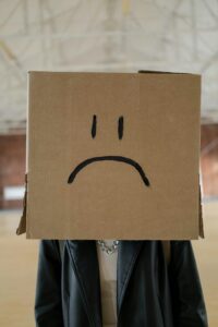 Person stands indoors with a cardboard box over their head, drawn-on sad face (two eyes and frowning mouth).