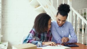 Two young women study together at a table, one guiding the other over an open notebook with a pen and tablet nearby.