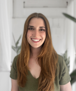 Smiling woman with long red hair wearing a green knit top, indoors with a blurred white background.