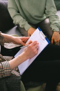 Person writes notes on a blue clipboard with a pen, while another person sits nearby in a green sweater.